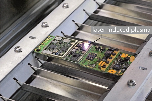 Printed circuit board of a mobile phone clamped in machine 5 while a laser spectroscopic measurement at an electronic component is conducted, see bright spot of laser-induced plasma.